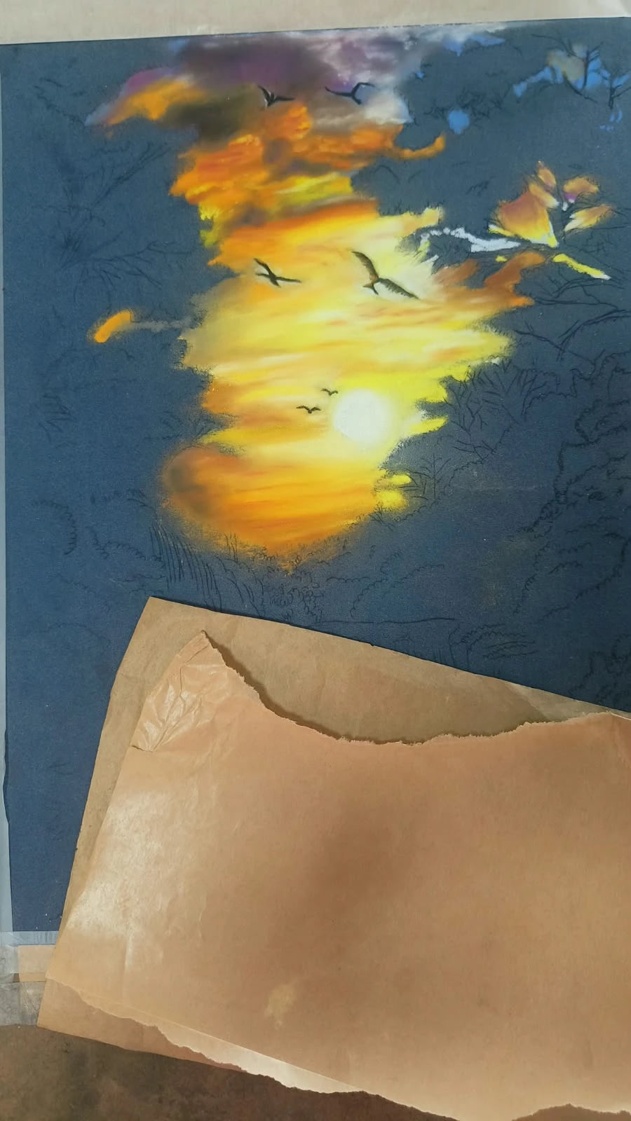 A soft pastel sunset in progress on dark blue paper: glowing orange clouds and birds are finished at the top, the trees below are still pencil lines, and brown paper protects the rest